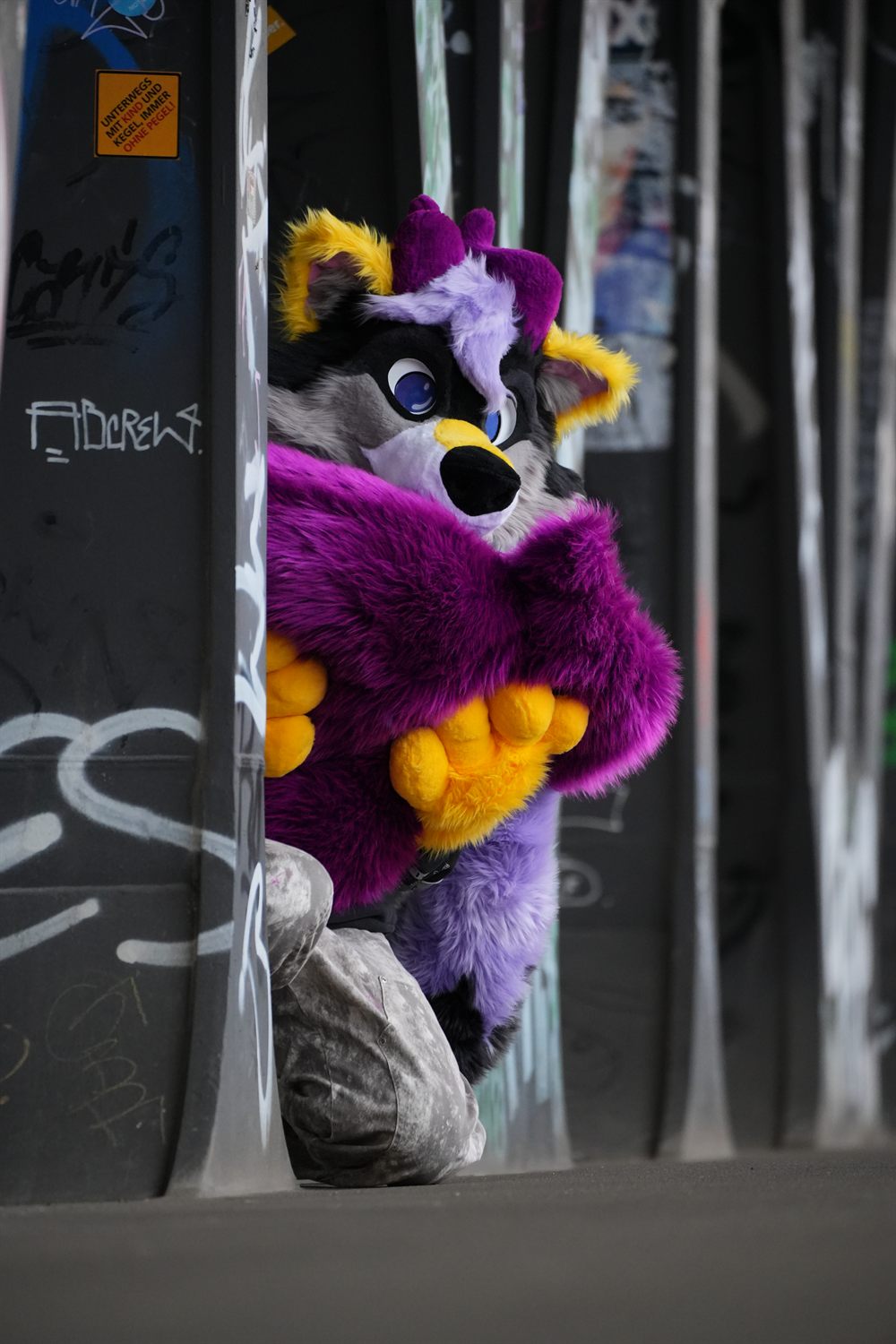 Fursuiter in Lila und Gelb schaut hinter einer Graffiti-Säule hervor